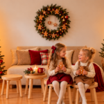 Zwei lachende Kinder mit Lebkuchen vor einem festlich geschmückten Weihnachtsbaum beim Weihnachts-Mini-Shooting von CoachOgrafie in Ismaning.